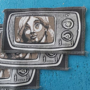 Könnte beinhalten: Mehrere rechteckige Aufnäher mit einem Vintage-Fernsehdesign. Jeder Aufnäher zeigt ein Graustufenbild eines menschlichen Gesichts auf dem Bildschirm, mit einem schwarzen Rand und weißen Nähten. Der Hintergrund ist einfarbig blau.
