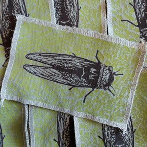 May include: Rectangular fabric patches with a lime green background and a repeating pattern of a dark brown cicada insect. Each patch has a white stitched border. The cicada illustration is detailed, showing wings and body features.