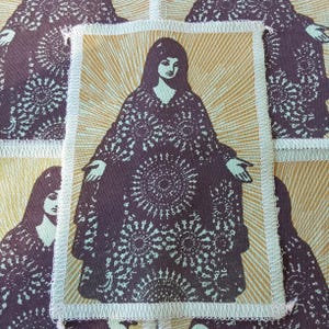 May include: A rectangular fabric patch featuring a screen-printed illustration of a figure with long hair wearing a dark purple robe adorned with circular starburst patterns, set against a light gold background with radiating sunburst lines.
