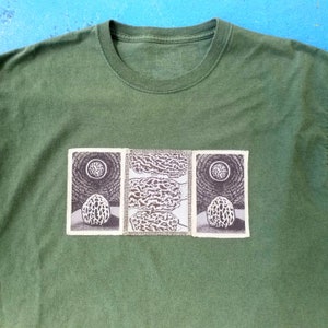 Könnte beinhalten: Ein olivgrünes T-Shirt mit drei rechteckigen Aufnähern auf der Brust. Die äußeren Aufnäher zeigen eine schwarz-weiße Illustration einer Morchel unter einem kreisförmigen Mond, während der mittlere Aufnäher ein strukturiertes Muster aufweist.