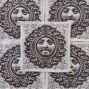 May include: A square fabric patch featuring a dark brown printed design of a human face centered within a circular frame, surrounded by intricate swirling patterns. The patch has visible stitched edges and is set against a matching repeating background.