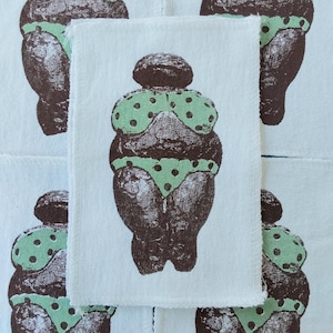 May include: A rectangular white fabric patch featuring a dark brown screen-printed graphic of a stylized female figure. The figure wears a light green bikini top and bottom with dark brown polka dots, set against a repeating pattern background.