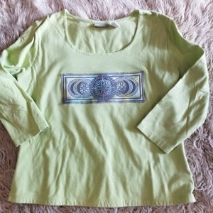 Op de afbeelding: Een lichtgroen shirt met lange mouwen met een blauw en groen grafisch ontwerp van een mandala met manen op een rechthoekige achtergrond.