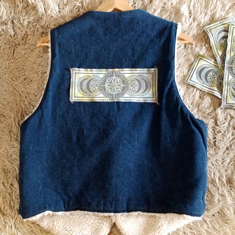 Op de afbeelding: Een blauwe denimvest met een cr&egrave;mekleurige fleecevoering. De vest heeft een rechthoekige patch met een geometrisch ontwerp in tinten geel, groen en bruin.