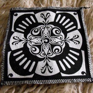 May include: Black and white fabric patch with a geometric design. The design features a central motif with four radiating arms, each with a series of curved lines. The patch has a black border with a white stitching detail.