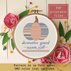 Può includere: Uno schema di ricamo a punto croce di una zucca decorativa con un design geometrico blu, arancione e bianco. Il testo "decorative gourd - season, y'all -" è sotto la zucca. Lo schema è a colori e include un elenco di colori DMC.