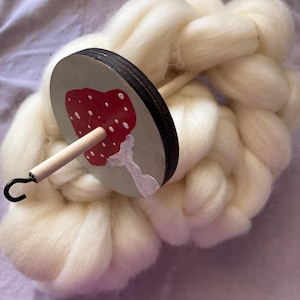 May include: A drop spindle featuring a circular wooden whorl painted with a red and white mushroom design, attached to a light-colored wooden shaft with a black metal hook at the tip, resting against a thick coil of soft, off-white unspun wool fiber.