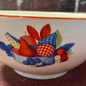 May include: White ceramic bowl with a red rim and a fruit design. The bowl features a blue and white basket filled with red, yellow, and blue fruits. The bowl is a classic kitchenware item.