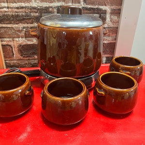 May include: A vintage brown fondue pot set with four matching bowls. The fondue pot has a silver lid and sits on a chrome base. The bowls have small handles. The set is on a red surface.