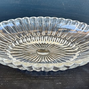 May include: Clear glass oval serving dish with a scalloped edge and ornate handles. The dish features a sunburst design in the center and is ideal for serving appetizers or desserts. The dish is made of clear glass.