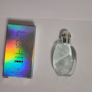 Könnte beinhalten: Eine durchsichtige Glasflasche mit Trussardi Light Him Parfum und einem silbernen Verschluss. Die Flasche steht auf einer weißen Oberfläche. Die Parfumverpackung befindet sich im Hintergrund und hat ein holografisches Design mit den aufgedruckten Worten "Trussardi" und "Light Him".