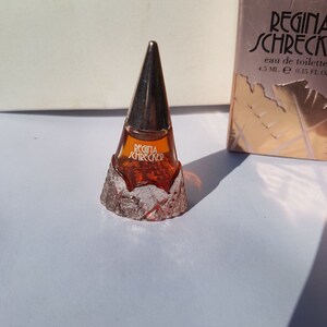 Puede incluir: Un pequeño frasco de perfume con una tapa cónica metálica y una base texturizada. El frasco contiene un líquido marrón dorado y tiene la inscripción "Regina Schrecker". El frasco es de 4,5 ml / 0,15 fl. oz.