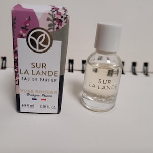 Op de afbeelding: Een kleine fles "Sur La Lande" Eau de Parfum en de bijbehorende doos. De doos is versierd met een bloemmotief in roze, paars en wit. De parfumfles is transparant met een witte dop. Inhoud: 5 ml.
