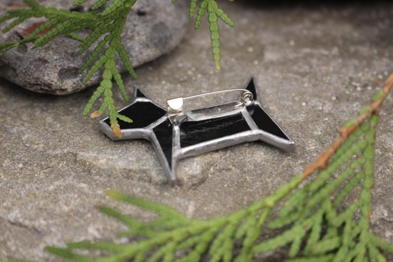 Pode incluir: Broche de vidro preto em forma de c&atilde;o com um alfinete de prata sobre um fundo de pedra cinza.
