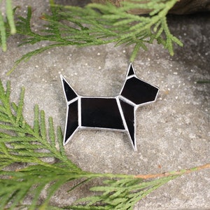 Pode incluir: Uma broche de vidro geom&eacute;trico preto em forma de c&atilde;o com detalhes prateados. A broche est&aacute; sobre uma superf&iacute;cie de pedra cinza com folhagem verde.