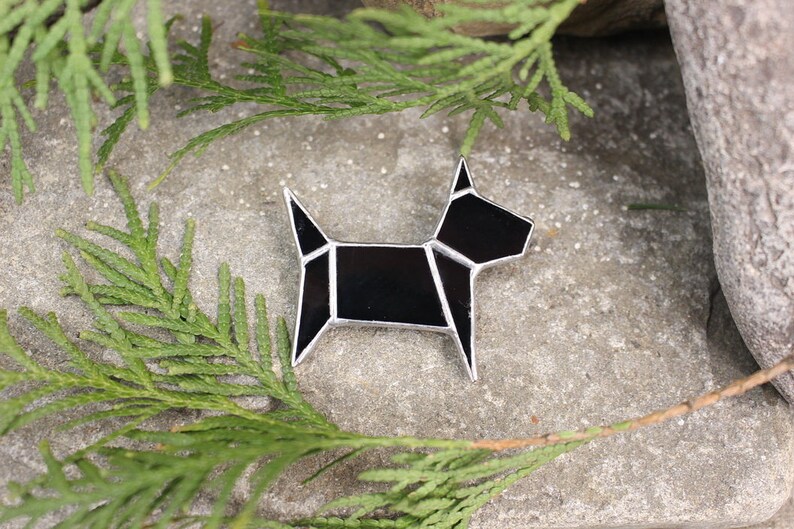 Pode incluir: Uma broche de vidro geom&eacute;trico preto em forma de c&atilde;o com detalhes prateados. A broche est&aacute; sobre uma superf&iacute;cie de pedra cinza com folhagem verde.