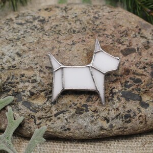 Op de afbeelding: Witte glazen hondenbroche met een geometrisch ontwerp. De broche ligt op een steen.