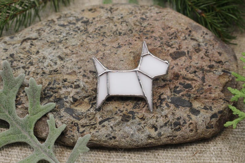 Op de afbeelding: Witte glazen hondenbroche met een geometrisch ontwerp. De broche ligt op een steen.