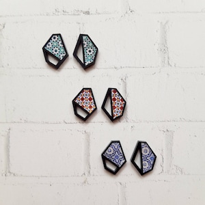 May include: Six geometric shaped earrings with black frames and colorful patterned inserts. The earrings are arranged in three pairs on a white brick wall.
