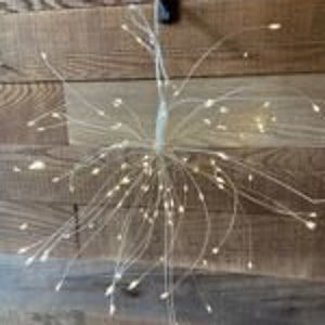 May include: A decorative light fixture with numerous small, warm white LED lights radiating outwards. The lights are attached to thin, flexible wires, creating a starburst effect. The fixture is hanging against a wooden wall.
