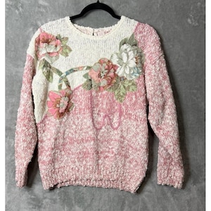 Op de afbeelding: Een vintage trui met een crèmekleurig bovenstuk en een roze en wit gespikkeld onderstuk. De trui heeft bloemenapplicaties in roze, wit en groen. De mouwen zijn lang en de halslijn is rond.