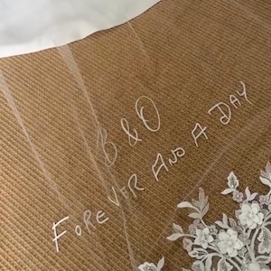 Pode incluir: Um véu de noiva branco transparente com detalhes de renda floral na borda inferior. O véu tem o texto manuscrito "B & O Forever And A Day" em escrita branca. O véu está sobre uma superfície marrom texturizada.
