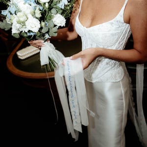 May include: A white lace corset top and satin skirt paired with a bouquet of blue and white flowers. A sheer white ribbon attached to the bouquet features the handwritten blue ink text, Congratulations Love Mum x, and a small heart symbol.