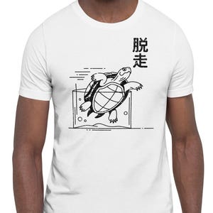 May include: White t-shirt featuring a black graphic of two turtles swimming in a rectangular shape. Above the turtles is Japanese text. The shirt is a crew neck style and is being worn.