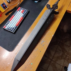 Puede incluir: Una espada grande de espuma gris con empuñadura y guarda de color negro, apoyada sobre una superficie de madera. La espada tiene una hoja larga y rectangular con tres cortes en forma de cruz. También se ven un ratón y un teclado de ordenador negros.