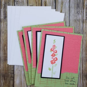 May include: Set of five blank greeting cards with a green and pink patterned design. The front of the card features a watercolour illustration of a pink flower with green leaves. The text "Live well, laugh often, love much" is written in black ink on the bottom right corner of the card.