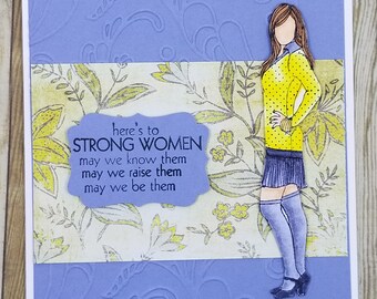 Puede incluir: Una tarjeta morada con un patrón floral y un recorte de una mujer con un suéter amarillo y una falda negra. La tarjeta tiene el texto "here's to STRONG WOMEN may we know them may we raise them may we be them".