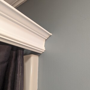 May include: White crown molding with decorative trim is installed at the top of a wall painted a muted blue-gray. A dark gray curtain is partially visible, adding contrast to the bright white molding.