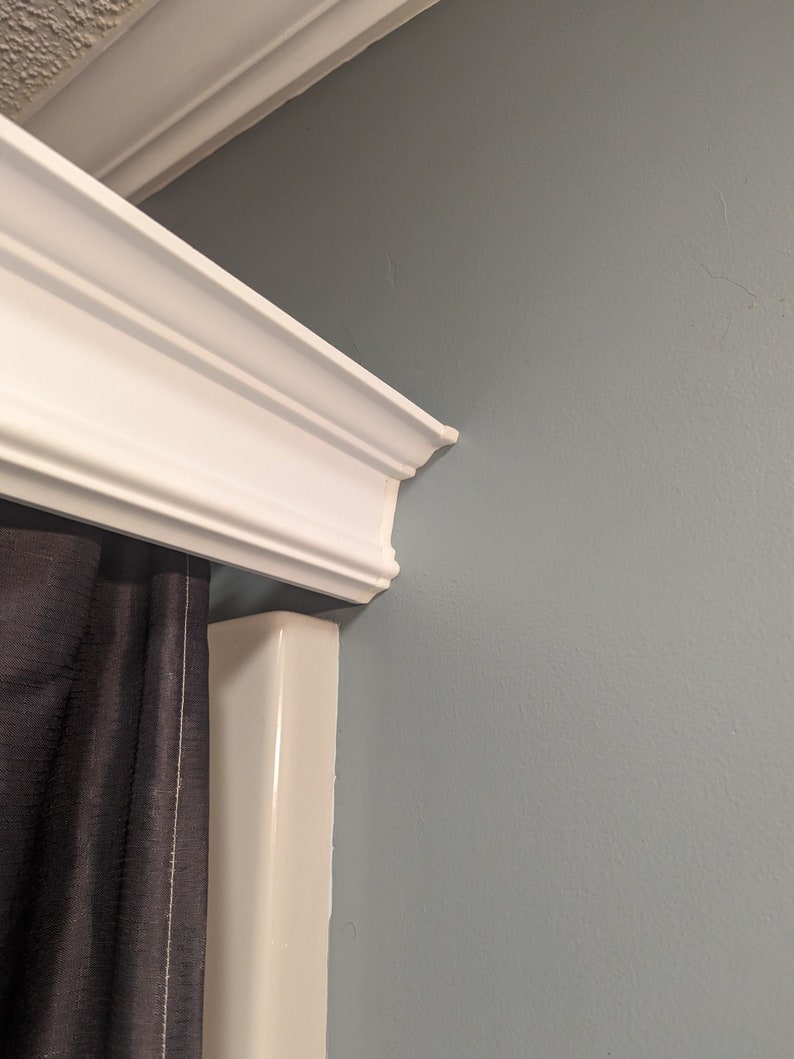 May include: White crown molding with decorative trim is installed at the top of a wall painted a muted blue-gray. A dark gray curtain is partially visible, adding contrast to the bright white molding.