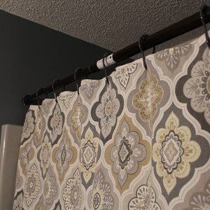 May include: A patterned shower curtain with a dark bronze rod and black hooks. The curtain features a repeating floral design in shades of gray, beige, and yellow. The curtain is hanging in a bathroom setting.