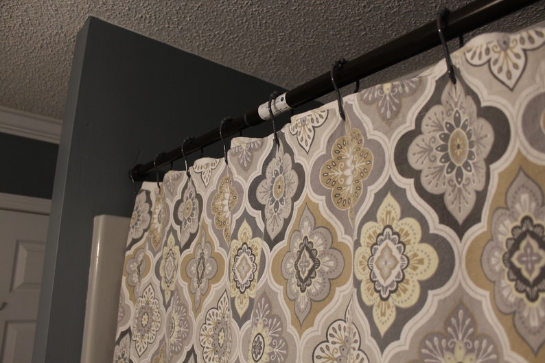 May include: A patterned shower curtain with a dark bronze rod and black hooks. The curtain features a repeating floral design in shades of gray, beige, and yellow. The curtain is hanging in a bathroom setting.