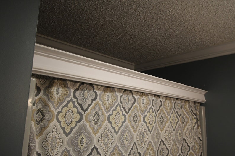 May include: White crown molding above a patterned curtain. The curtain features a repeating design in gray, yellow, and white. The ceiling is a textured brown color, and the wall is a dark gray.