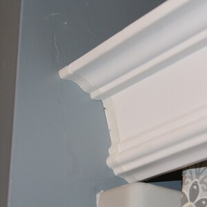 May include: A close-up of a white crown molding detail. The molding is installed on a light blue wall. The corner of a gray wall is visible on the left side of the image.