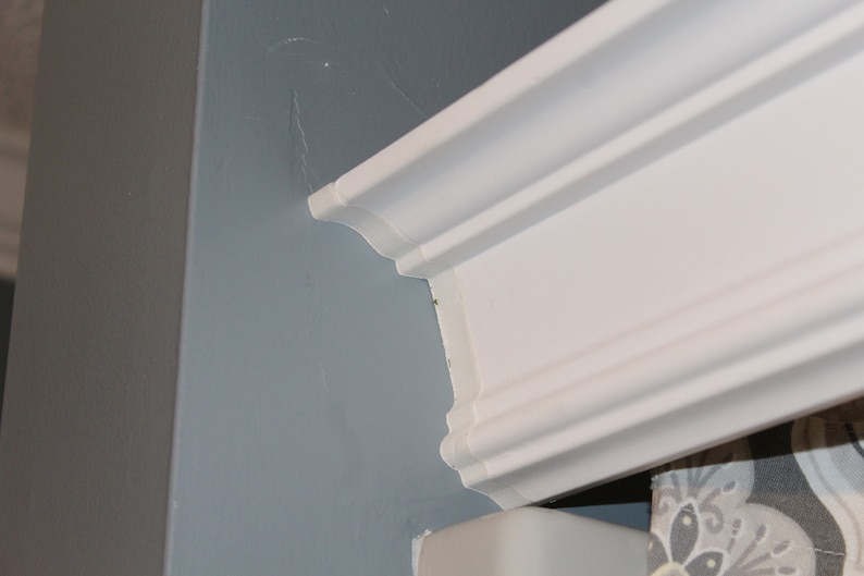 May include: A close-up of a white crown molding detail. The molding is installed on a light blue wall. The corner of a gray wall is visible on the left side of the image.