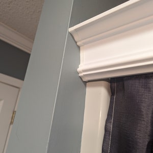 May include: A corner view of a room featuring a white crown molding and a light blue wall. A dark gray curtain is partially visible. The ceiling is off-white. The molding is a decorative element.