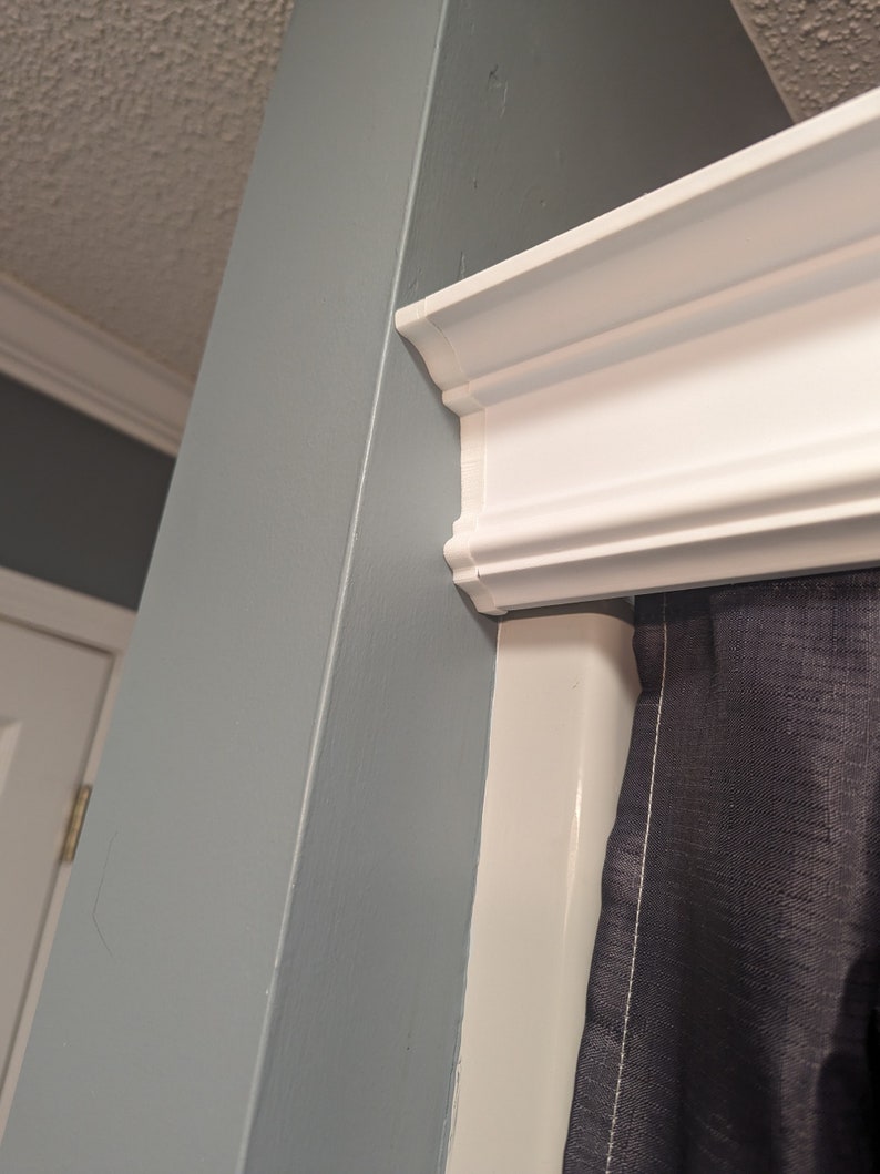 May include: A corner view of a room featuring a white crown molding and a light blue wall. A dark gray curtain is partially visible. The ceiling is off-white. The molding is a decorative element.
