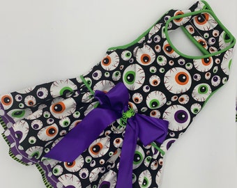 May include: A sleeveless dress featuring a black background covered in white, green, and orange eyeball designs. The dress has a purple bow and green trim. The hemline and neckline are trimmed with green and purple striped fabric.