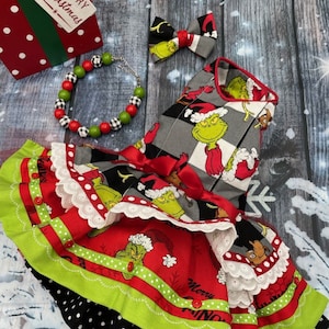 May include: A pet dress featuring a Grinch print on a grey and black checkered bodice and a red skirt with white lace trim, green ruffles, and red buttons. Includes a matching bow tie and a beaded necklace with red, green, and black-and-white beads.