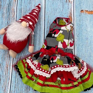 May include: A gnome with a red and white striped hat and a white beard next to a dress featuring a Grinch print on a grey and black buffalo plaid bodice, a red skirt with green trim, white lace accents, and a red ribbon bow at the waist.