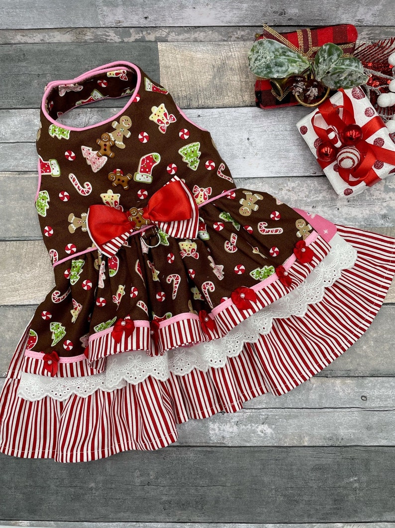 May include: A brown pet dress featuring a gingerbread man, candy cane, and Christmas tree print. The bodice has a red satin bow with a small gingerbread charm. The skirt has tiered layers of red and white striped fabric and white eyelet lace.