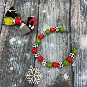 May include: A fabric bow tie featuring a green character print on a black background and a beaded bracelet with alternating red, green, and black and white buffalo plaid beads, connected by a silver-toned metal chain with a lobster claw clasp.