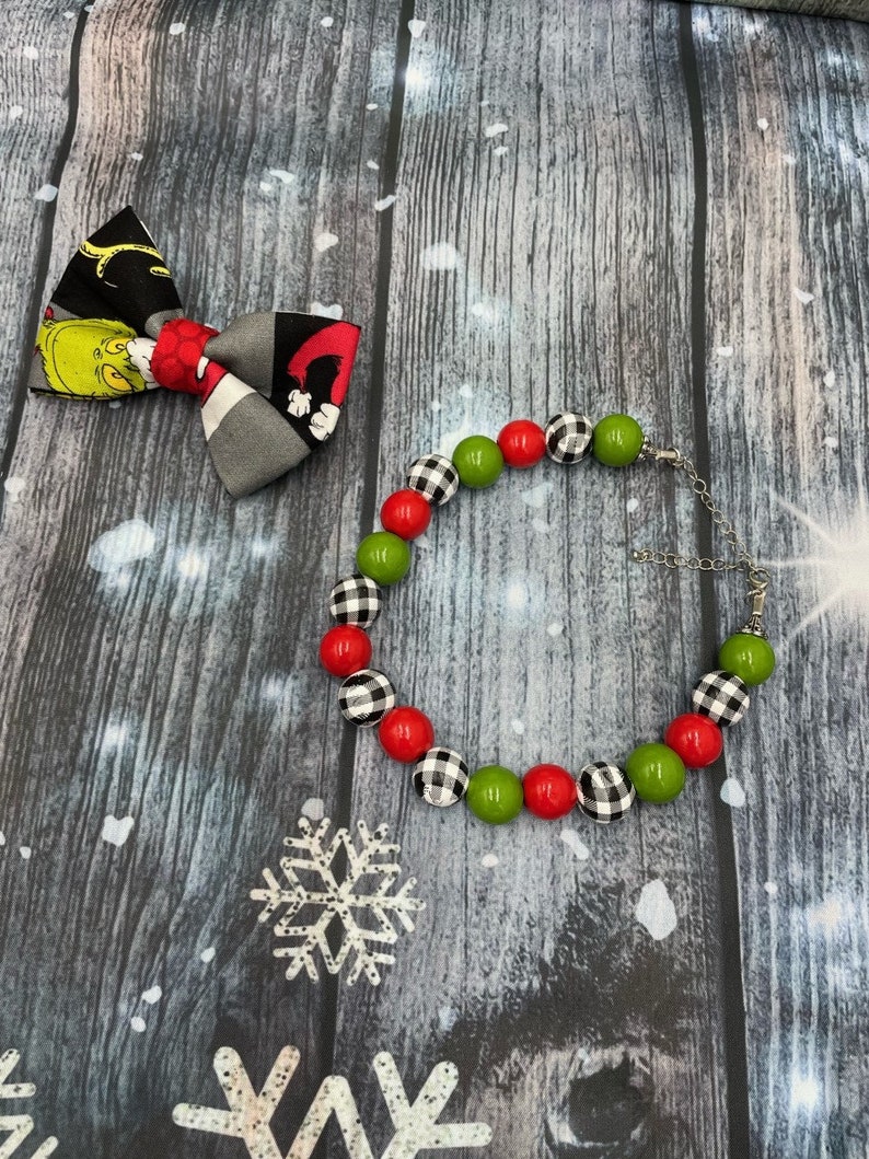 May include: A fabric bow tie featuring a green character print on a black background and a beaded bracelet with alternating red, green, and black and white buffalo plaid beads, connected by a silver-toned metal chain with a lobster claw clasp.
