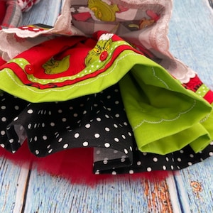 May include: A multi-layered skirt featuring a red fabric with a green character print, a black fabric with white polka dots, and a solid lime green layer. The skirt includes a white scalloped lace trim at the top and a red tulle layer at the bottom.