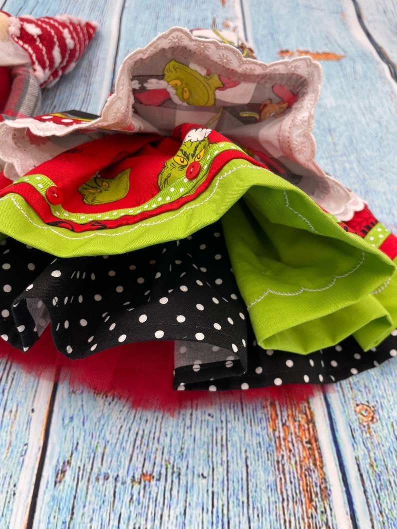 May include: A multi-layered skirt featuring a red fabric with a green character print, a black fabric with white polka dots, and a solid lime green layer. The skirt includes a white scalloped lace trim at the top and a red tulle layer at the bottom.