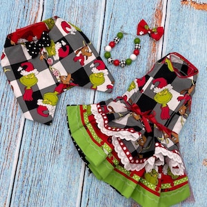 May include: A pet outfit set featuring a black and white buffalo plaid fabric with a Grinch character print. The set includes a vest with a black polka dot bow tie, a ruffled dress with red ribbon and white lace trim, and a beaded collar with a bow.