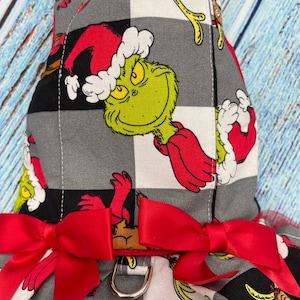 May include: A pet harness featuring a grey and black checkered pattern with a repeating print of a green character wearing a red Santa hat. The harness includes red fabric trim, two red satin bows at the front, and a silver metal D-ring.